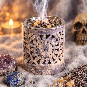 Könnte beinhalten: Ein zylindrischer Räuchergefäß aus Stein mit kunstvollen floralen Ausschnitten und einem Metallgitter oben. Weiße Rauchschwaden steigen aus der Mitte auf, die eine Mischung aus getrockneten Lavendelblüten und Harz-Räucherwerk enthält.