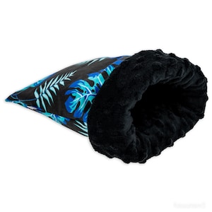 Puede incluir: Una cama para mascotas de color negro y azul con estampado tropical y un forro de felpa negro suave. La cama tiene forma de cilindro largo y estrecho.