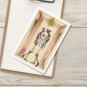 Puede incluir: Una impresión que muestra dos esqueletos abrazándose, con un corazón rojo entre ellos, bajo un muérdago. Los esqueletos están frente a una cortina. La impresión está en una tarjeta blanca, sobre una carpeta.