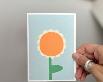 Könnte beinhalten: Eine Postkarte mit einer einfachen Illustration einer Blume mit weißem Umriss, orangefarbenem Zentrum und grünem Stiel auf hellblauem Hintergrund.