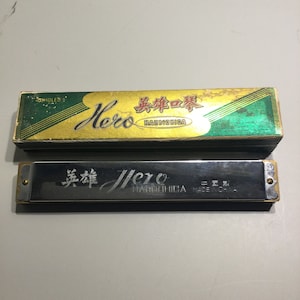 May include: A silver harmonica with the word "Hero" and Chinese characters printed on the front. The harmonica is in its original box, which is green and gold with the word "Hero" and Chinese characters printed on it.