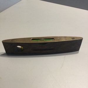 Puede incluir: Un nivel de latón con una base de madera. El nivel tiene una burbuja verde y el texto "SAMUELSON & CO. NO.1025 CORRECT" está impreso en el latón.