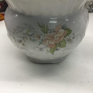 Puede incluir: Un macetero de cerámica blanca con un borde festoneado y un borde dorado. El macetero presenta un diseño floral con flores rosas, blancas y verdes.