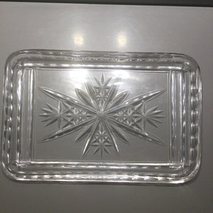 Puede incluir: Una bandeja de servir de vidrio transparente con un patrón de estrella decorativo grabado en la parte inferior. La bandeja tiene un borde elevado y un borde ligeramente redondeado.