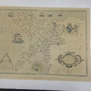 May include: An antique map of Caernarvonshire and Anglesey, Wales, with a detailed depiction of the region's geography and towns. The map is printed on aged paper with a cream background and features intricate details in brown, black, and blue ink.