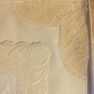 May include: A white linen handkerchief with cream-coloured lace detailing. The lace features floral and geometric patterns around the edges. Presented in a white box with a decorative lid.