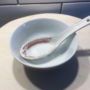 Puede incluir: Un cuenco de cerámica blanco con una cuchara a juego. El cuenco tiene un borde rojo y dorado, y la cuchara presenta un diseño floral. La cuchara descansa dentro del cuenco, creando una disposición de vajilla clásica.