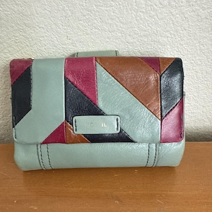 May include: A bi-fold wallet featuring a geometric patchwork design in shades of sage green, deep magenta, brown, and black leather. The front flap displays a rectangular leather patch embossed with the Fossil brand name in metallic lettering.