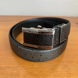 May include: A rolled-up gray belt with a textured pattern and a black buckle. The buckle has a silver frame and the word "RONZ" is visible. The belt is on a wooden surface.