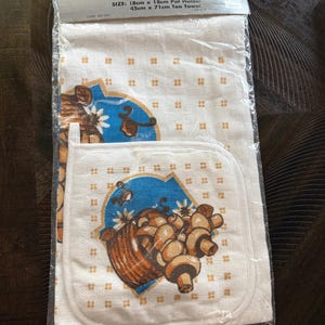 May include: A 2-piece kitchen set includes a pot holder and a tea towel. The white fabric features a repeating pattern of small orange squares. The pot holder and towel have a design of a basket of mushrooms with a blue and white background. The pot holder is 7 inches by 7 inches.