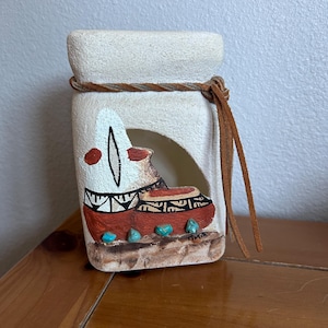 May include: A cream-colored, rectangular candle holder with a Native American design. The design features a red and brown pot with turquoise accents. A leather cord wraps around the top, and a leather strip hangs on the side.