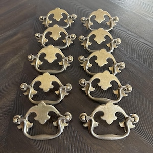 May include: A set of ten matching brass drawer pulls arranged in two columns. Each pull features a decorative backplate with a central leaf-like motif and a curved, swinging handle attached to two side posts, showcasing a metallic gold finish.