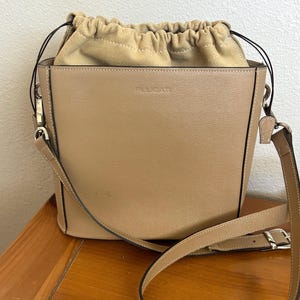 May include: A tan leather bucket bag with a long strap and a drawstring closure. The bag has a small silver logo that says "Furla" on the front.