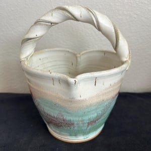 May include: Handmade ceramic basket with a twisted handle. The basket features a white, teal, and brown color scheme. The handle is white with a twisted design. The basket has a unique shape and is suitable for decorative use.