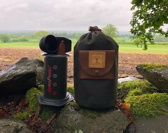 Può includere: Una macchina da caffè AeroPress nera con una custodia in pelle marrone. La custodia ha un logo ad albero sul davanti.