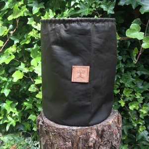 Könnte beinhalten: Eine dunkelgrüne zylindrische Tasche mit Kordelzugverschluss. Ein kleines, quadratisches, braunes Lederpatch mit einem Baumdesign ist auf der Vorderseite aufgenäht. Die Tasche steht auf einem Baumstumpf, mit grünem Laub im Hintergrund.