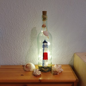 Könnte beinhalten: Eine klare Glasflasche mit einem Korkverschluss und einem gemalten Leuchtturmdesign im Inneren. Die Flasche wird von innen mit weißen Lichterketten beleuchtet und ist von Muscheln auf einer Holzoberfläche umgeben.