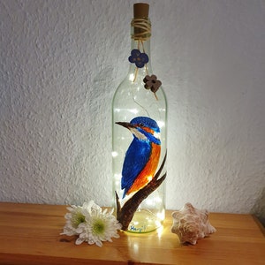 Könnte beinhalten: Eine klare Glasflasche mit einem Korkverschluss und einem gemalten Bild eines blauen und orangefarbenen Eisvogels, der auf einem Ast sitzt. Die Flasche ist mit weißen Lichterketten beleuchtet und mit kleinen Holzblumen verziert.