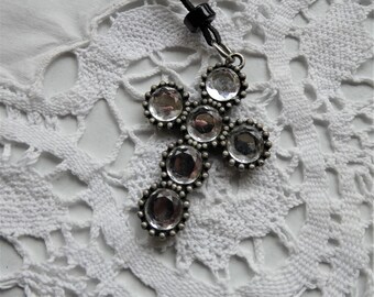 Peut inclure: Un pendentif en forme de croix argentée avec des strass transparents, suspendu à un cordon noir.