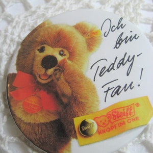Könnte beinhalten: Ein runder, weißer Knopf mit einem braunen Teddybär, der eine rote Schleife trägt. Der Knopf trägt den Text "Ich bin Teddy Fan!" und ein gelbes und rotes Steiff-Logo mit dem Text "Knopf im Ohr".
