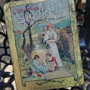 Op de afbeelding: Een vintage blikken doos met een gouden achtergrond en een vervaagde afbeelding van drie kinderen. De doos heeft de tekst "Chocolat Suchard" en "Collection 'Les Affiches Suchard'" in gouden letters.