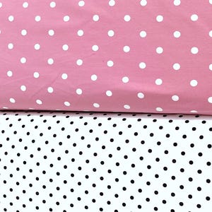 Könnte beinhalten: Zwei Stoffrollen sind horizontal gestapelt. Die obere Rolle besteht aus einfarbigem rosa Stoff mit einem sich wiederholenden Muster aus großen weißen Punkten. Die untere Rolle besteht aus weißem Stoff mit einem dichten Muster aus kleinen schwarzen Punkten.