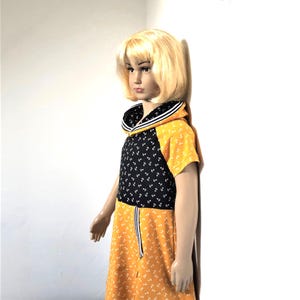 May include: A yellow and black dress with a white anchor print. The dress has a sailor collar and a hood. The dress is on a mannequin with blonde hair.