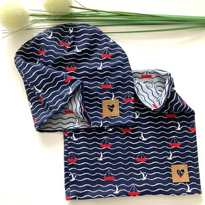 Peut inclure: Un ensemble bonnet et tour de cou bleu et blanc à motifs avec des voiliers rouges et des oiseaux blancs. Le tissu est orné d'un patch en cuir marron avec une découpe en forme de cœur.