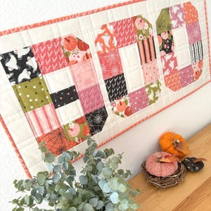 Könnte beinhalten: Ein Wandbehang im Patchwork-Stil mit dem Wort "BOO" zum Thema Halloween. Die Buchstaben zeigen verschiedene Muster, darunter Geister, Kürbisse und Spinnennetze, in Orange, Rosa, Schwarz und Grün. Das Ganze ist mit einem korallenfarbenen Stoff eingefasst.