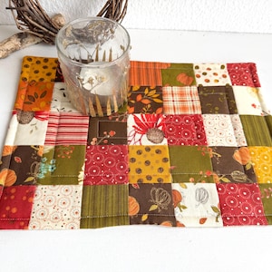 Op de afbeelding: Een patchwork tafelloper met herfststoffen in rood, oranje, groen en bruin. Een heldere glazen kandelaar met een brandende kaars staat erop. De loper is ongeveer 30 cm breed en 46 cm lang.