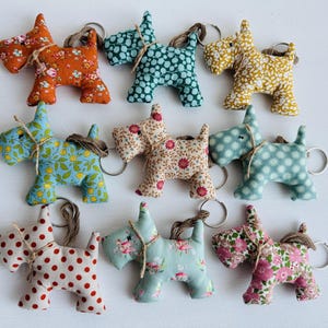Op de afbeelding: Een verzameling handgemaakte stoffen honden sleutelhangers in verschillende kleuren en bloempatronen. Elke sleutelhanger heeft een metalen ring en een touwdetail. De honden zijn in tinten oranje, blauw, geel en roze.