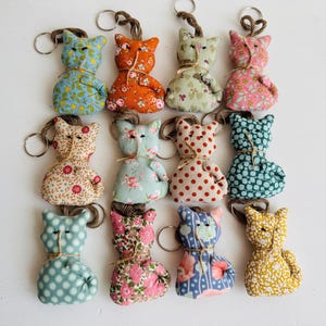 Op de afbeelding: Een verzameling handgemaakte katvormige sleutelhangers. Elke sleutelhanger is gemaakt van stof met patronen in verschillende kleuren, waaronder blauw, oranje, roze en geel. De sleutelhangers hebben een eenvoudig ontwerp met een kleine lus om sleutels aan te bevestigen.