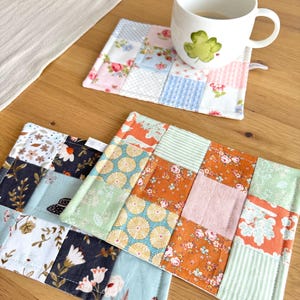 Könnte beinhalten: Drei rechteckige Stoffuntersetzer mit Patchwork-Designs. Ein Untersetzer zeigt florale Pastellmuster und Vichy-Karos mit einer weißen Tasse. Zwei weitere Untersetzer bestehen aus dunkelblauen, orangen und hellgrünen floralen Stoffquadraten.