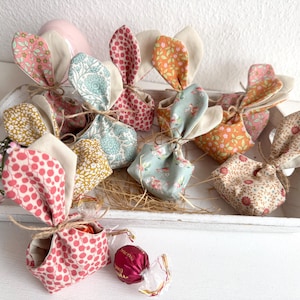 Könnte beinhalten: Eine Sammlung von stofflichen Geschenkbeuteln in Hasenform mit verschiedenen floralen Mustern, darunter Rosa, Orange und Blau, auf einem weißen Tablett angeordnet. Jeder Beutel ist mit Bindfaden gebunden und hat beige Stoffohren. Ein rosa Ei und rosa Blumen sind ebenfalls vorhanden.