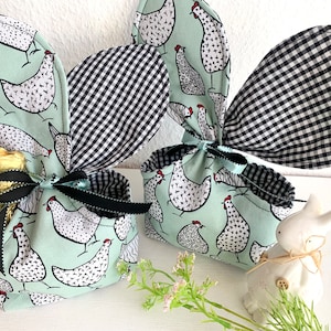 Op de afbeelding: Twee paasgeschenktassen met konijnenooraanzetten. De tassen zijn lichtgroen met een zwart-wit kippenpatroon. De konijnenoren zijn zwart-wit geruit. Elke tas is vastgebonden met een zwart lint. Een goudfolie chocoladekonijn is zichtbaar.