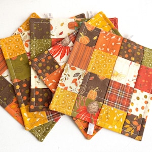 Peut inclure: Quatre maniques en tissu patchwork dans des tons de brun, orange, jaune et blanc. Les maniques sont composées de carrés de tissus différents avec divers motifs, notamment du plaid, des fleurs et des motifs géométriques.