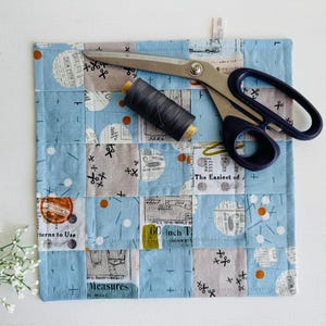 Peut inclure: Un tapis de couture en patchwork avec un motif bleu et gris, avec des ciseaux et une bobine de fil noir. Le tapis porte les inscriptions "Patterns to Use" et "60 inch T".