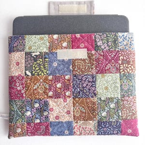 Peut inclure: Une housse pour ordinateur portable matelassée avec un motif patchwork. La housse présente divers motifs floraux dans les tons rose, bleu, vert et marron. Elle a un dessus gris et une fermeture velcro beige.
