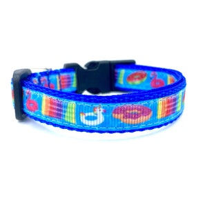 May include: A blue dog collar with a repeating pattern of colorful pool floats, including donuts and flamingos, on a light blue background. The collar has a black plastic buckle and a blue trim.