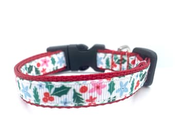 May include: A festive pet collar with a white background, adorned with a repeating pattern of pink and blue flowers, green leaves, and red berries. The collar has a red trim, a black plastic buckle, and a small silver bell.