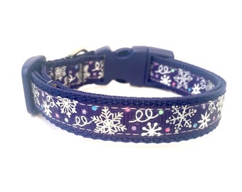 May include: A navy blue dog collar with a winter theme. The collar features a repeating pattern of white snowflakes and swirls on a dark blue background. The collar has a navy blue plastic buckle and a matching adjustable strap.