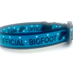 May include: A blue and black pet collar with the words "OFFICIAL BIGFOOT TEAM" in white lettering. The collar features a Bigfoot footprint graphic and white diamond shapes. The collar is made of a durable material and has a black plastic buckle.