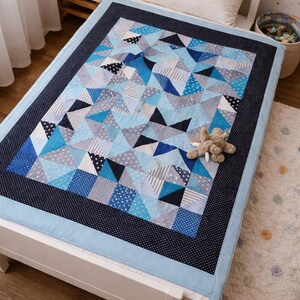 Könnte beinhalten: Eine handgefertigte Steppdecke mit einem geometrischen Muster in Blau-, Grau- und Weißtönen. Die Steppdecke zeigt Dreiecke, Quadrate und Punkte, eingerahmt von einem dunkelblauen Rand mit weißen Punkten. Ein kleines Stofftier liegt auf der Steppdecke.