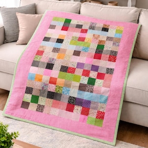 Könnte beinhalten: Eine Patchwork-Decke mit rosa Rand und grün gestreiftem Rand liegt auf einem beigen Sofa. Die Decke besteht aus verschiedenen bunten Stoffquadraten, darunter Blumen-, Tupfen- und unifarbene Muster. Ein gemütliches Wohnaccessoire.