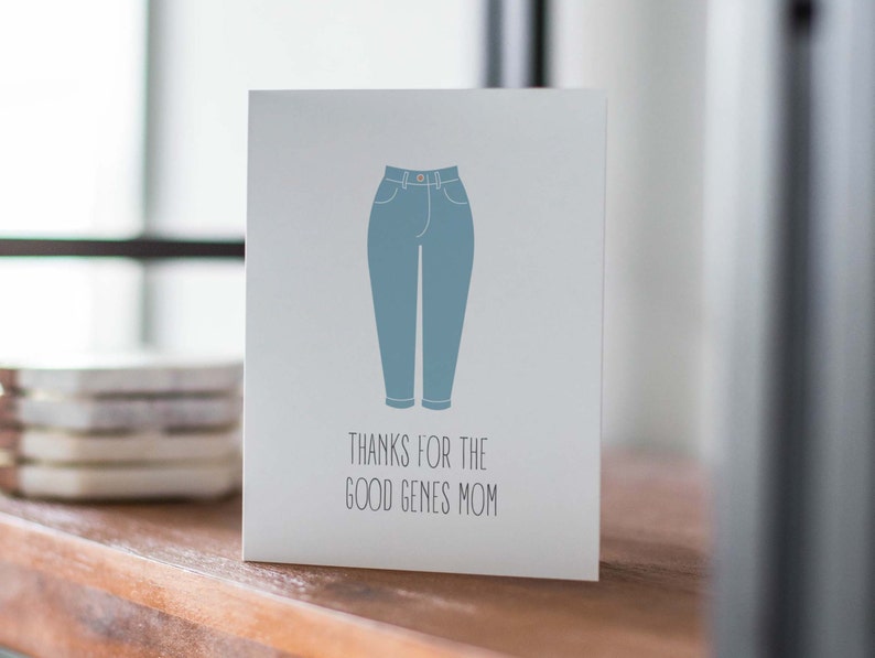 White card with illustration of mom jeans with "thanks for the good genes mom" written below. Lifestyle photo of card on shelf.