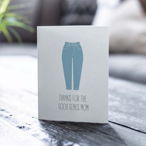 White card with illustration of mom jeans with "thanks for the good genes mom" written below. Lifestyle photo of card on table.