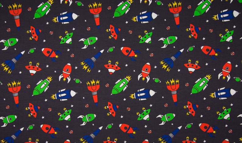 May include: A dark blue fabric with a repeating pattern of colorful rockets, spaceships, and planets. The rockets are red, green, and blue with yellow flames. The spaceships are blue with white windows. The planets are orange, yellow, and green.