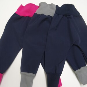 Könnte beinhalten: Vier Paar marineblaue Hosen mit farbigen Bündchen und Bund. Ein Paar hat ein leuchtend pinkfarbenes Bündchen und einen Bund, ein anderes hat ein graues Bündchen und einen Bund, und die anderen beiden haben marineblaue und graue Bündchen und Bünde. Die Hosen sind aus weichem Material.