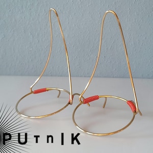 Op de afbeelding: Twee goudkleurige metalen standaards met een cirkelvormige basis en een gebogen bovenkant. De standaards hebben rode gewikkelde accenten. De standaards zijn waarschijnlijk bedoeld om items te presenteren. De achtergrond is een lichtblauwe muur.