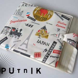 Puede incluir: Un álbum de viaje de vinilo blanco con un diseño colorido que presenta ilustraciones de monumentos, aviones y trenes. El álbum tiene una cubierta de plástico transparente y una correa blanca con cierre de botón. El texto en el álbum incluye "JUMBO", "SAS", "LUFTHANSA", "JAPAN", "Teneriffa", "Sizilien Spanien", "Jugoslawien" y "Garie".